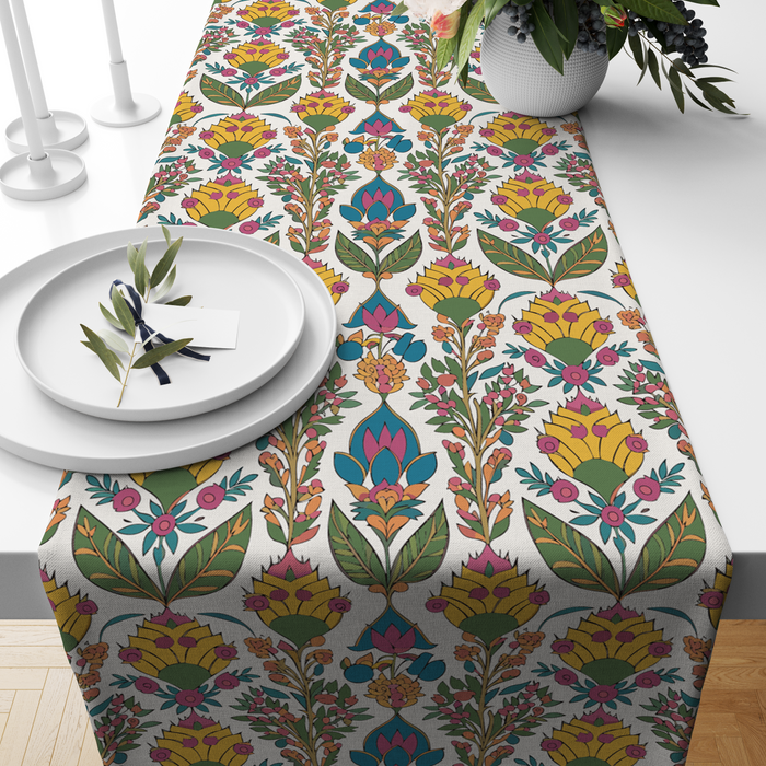 Cream table runner with a bold yellow blossom and green leaf print, pink and teal accents, styled on a wooden dining table
