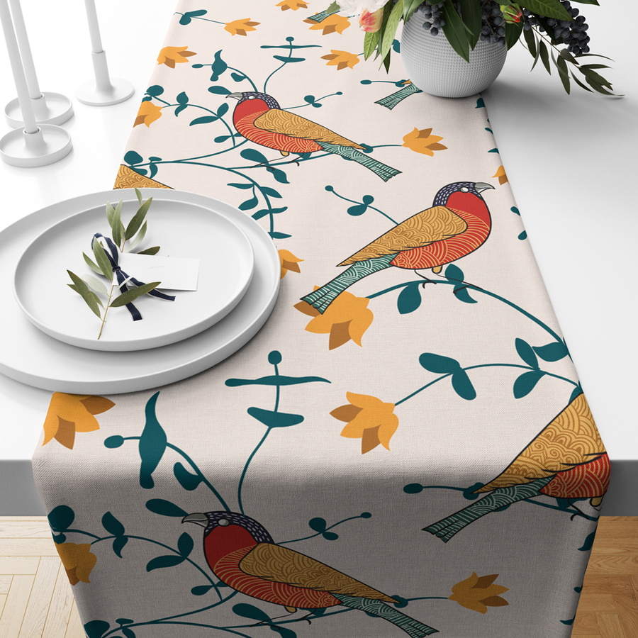 Cream table runner with a red and gold bird print among teal leaves and yellow flowers, on a dining table