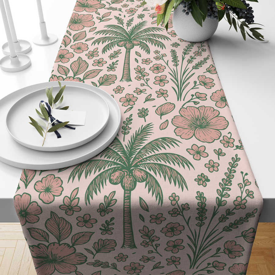 Blush-pink table runner with sage-green palm trees and floral motifs, styled on a wooden dining table