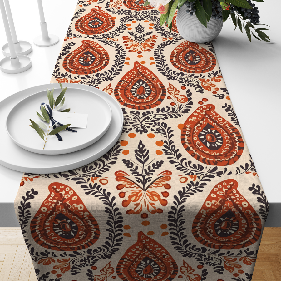 Cream table runner with a rust-brown paisley teardrop motif and navy leaf detailing, styled on a wooden dining table