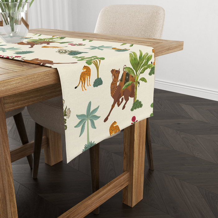 Cream table runner with camel, leopard, and palm-tree motifs, shown from another angle on a wooden table with tableware