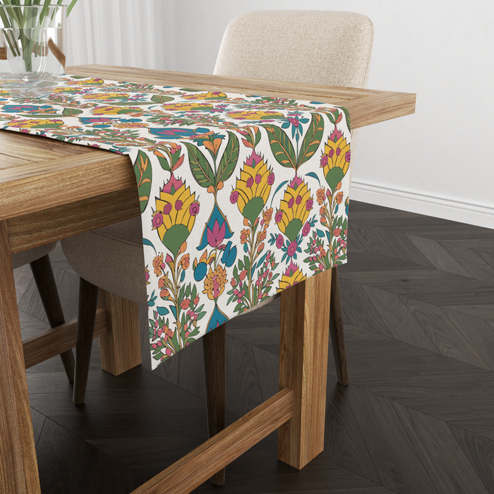 Cream table runner with a bold yellow blossom and green leaf print, pink and teal accents, shown from another angle on a wooden table with tableware